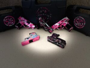 A collection of customized guns including Sig Sauer P365s, P320s, and Ruger LCP .380 pistols displayed alongside matching Sig Sauer cases that complement their unique colors and custom pink gun patterns.